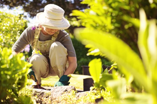 A woman wearing a wide-brimmed straw hat, beige gardening apron, and gloves is kneeling on a neatly maintained lawn in a garden, actively planting or tending to small plants or flowers. The garden features a lush, green lawn with a slightly textured grass surface, bordered by well-defined flower beds and hedges. In the background, there are tall trees and shrubs, providing natural privacy and shade, with sunlight filtering through the foliage and casting gentle shadows across the scene. The garden appears to be part of a residential outdoor space in Teddington, with a peaceful and well-maintained environment indicative of professional gardening or landscaping services. The soil in the flower bed looks rich and dark, contrasting with the vibrant foliage, and garden tools or pots are partially visible nearby. Overall, the scene exemplifies careful garden maintenance, suitable for clients seeking lawn care, planting, and outdoor landscaping in the Teddington area, as offered by Gardening Services Teddington.