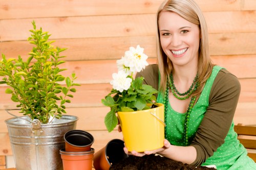 A woman with shoulder-length blonde hair and a bright smile is sitting outdoors in a garden setting, holding a yellow flowerpot with a flowering plant that has white blooms and green foliage. She is wearing a green sleeveless top with a beaded necklace and an olive-green cardigan. In front of her on a wooden surface are several empty flowerpots, including a small black one and a terracotta one. Behind her, there is a wooden fence, suggesting a private garden space, with a potted shrub in a metal container to her left. The garden layout features a mix of lush green lawn, bordered by a flower bed or planting area, with visible paving stones nearby. The environment appears well-maintained and cared for, with natural daylight illuminating the scene, indicating a bright and clear day. The overall setting showcases typical garden elements suitable for outdoor lawn and plant care, reflecting services provided by Gardening Services Teddington in the local TW postcodes.