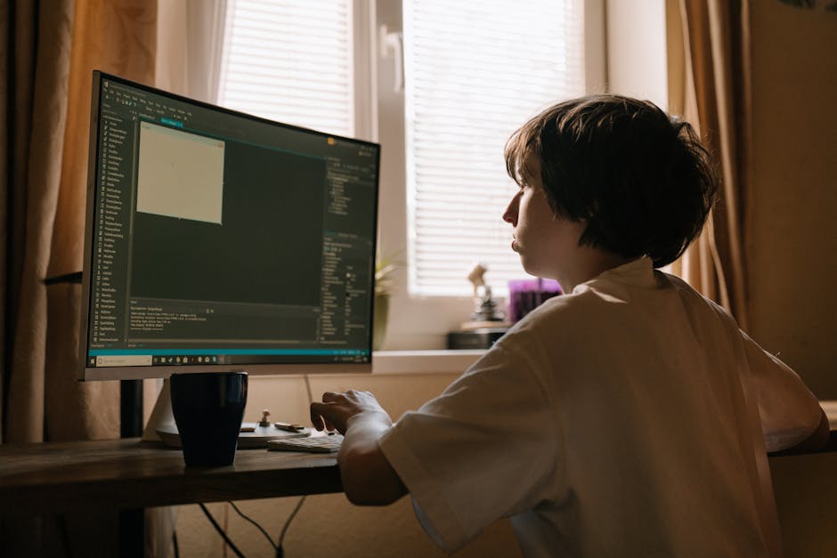 A woman with short, dark hair is seated at a wooden desk in a room with beige walls and a large window with partially open blinds, allowing natural light to fill the space. She is focused on a large c