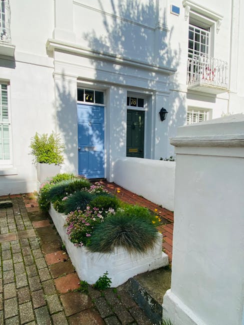 The front garden of a white townhouse features a neatly maintained flower bed bordered by white walls and a small brick pathway. The flower bed contains an assortment of low, rounded evergreen shrubs 