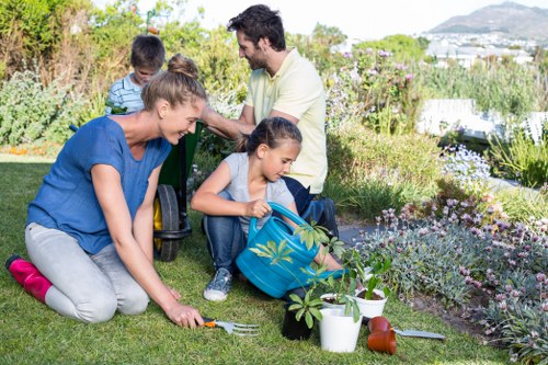 A family of four working together in a lush backyard garden, with two children, a girl and a boy, engaged in planting or watering young plants, while two adults, a woman and a man, supervise and assist. The garden features a well-maintained green lawn, flower beds filled with a variety of blooming plants and shrubs, and garden tools and pots scattered around for planting. In the background, there are mature trees, dense shrubbery, and a view of distant hills or water, indicating an outdoor space suited for gardening and outdoor activities. The scene is captured on a bright, sunny day with natural daylight highlighting the vibrant colours of the garden, showcasing healthy plants and well-kept lawn, illustrating a typical family gardening day that aligns with outdoor maintenance and landscaping services in Teddington, close to postcode TW11, provided by Gardening Services Teddington.