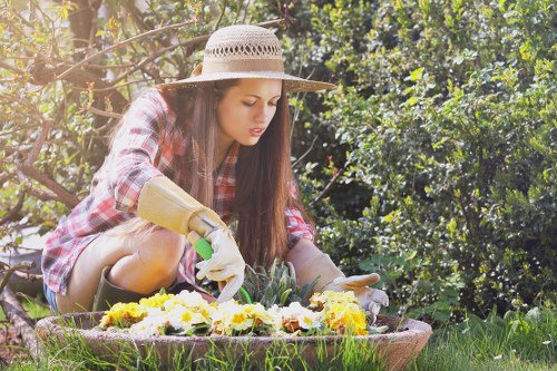 A young woman dressed in a plaid shirt, wearing a straw sun hat and gardening gloves, is kneeling on the grass in a lush garden in Teddington. She is tending to a circular flower bed that is filled with blooming plants displaying a mix of yellow, white, and purple flowers. The garden features a well-maintained lawn with dense, green shrubs and trees in the background, providing natural shade and enclosure. The scene suggests an outdoor space actively cared for, with healthy, vibrant plant life, indicative of professional gardening services in the Teddington area as offered by Gardening Services Teddington, highlighting a typical landscaped garden in a suburban setting under bright, natural daylight conditions.